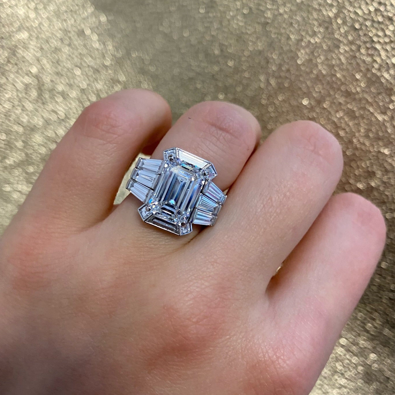 A hand displays the Bespoke Geometric Bezel Set Baguette and Asscher Cut Diamond Ring Jacket in white gold, set against a glittery gold background.