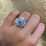 A hand wears the Geometric Bezel Set Baguette and Asscher Cut Diamond Ring Jacket in White Gold, featuring a rectangular design, against a glittery gold background.