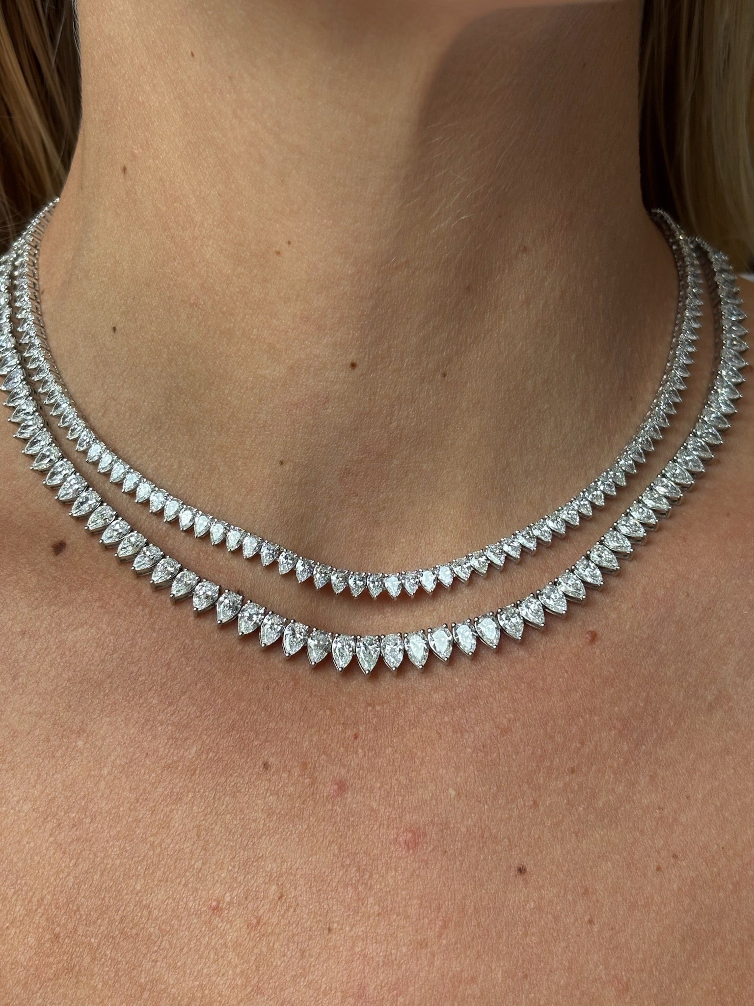 A close-up of a woman's neck wearing two layered necklaces: the upper is the Pear Diamond Tennis Necklace in White Gold, while the lower features larger marquise-shaped diamonds.