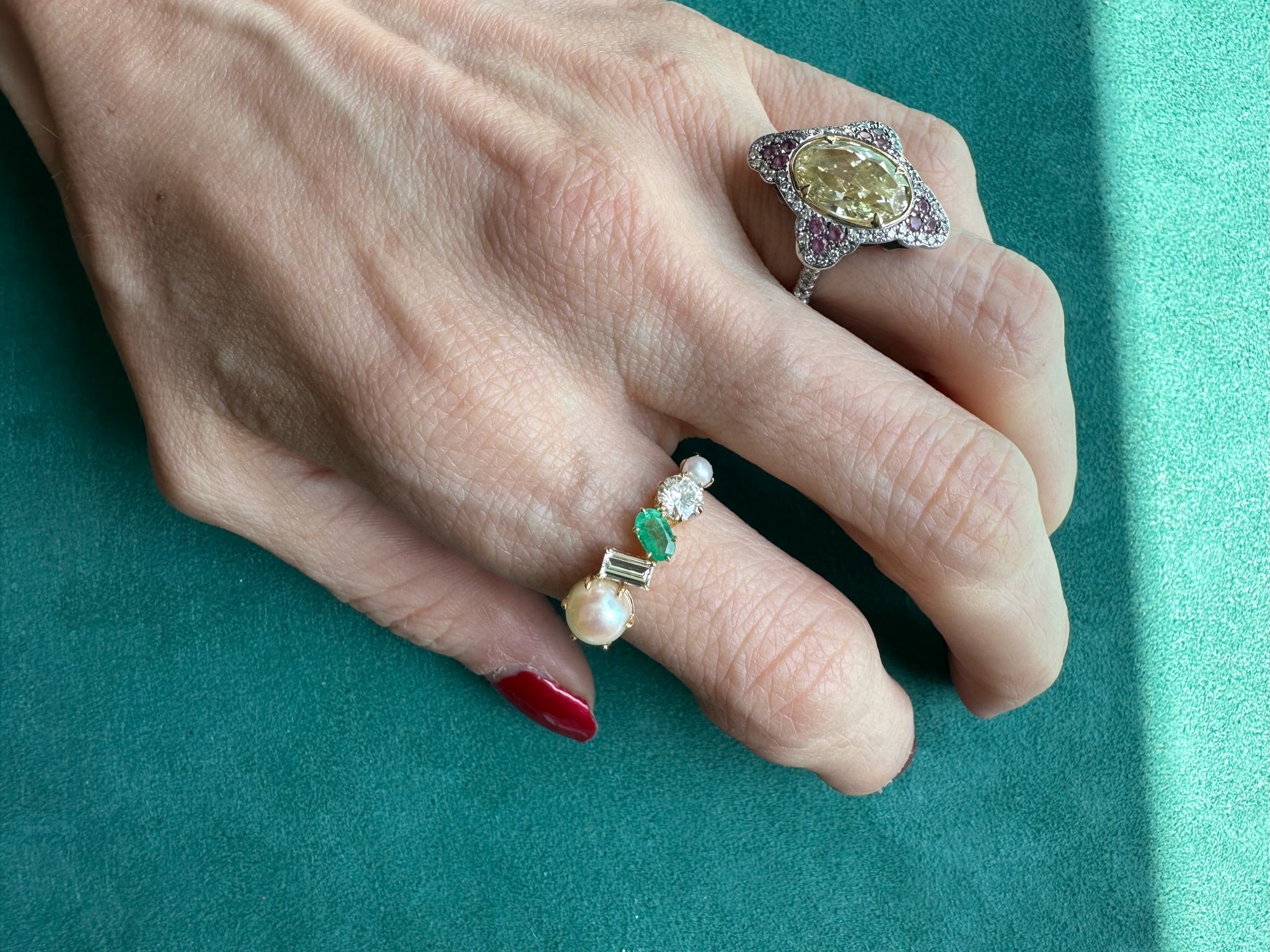 A hand with red nail polish wears two rings: a large yellow gemstone celebration ring and the Mixed Shape Pearl, Diamond and Emerald Half-Eternity Band, both shown on a teal surface.