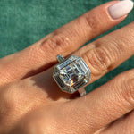A hand with light skin tone wears the 1980s Diamond Ring, Mounted by Oscar Heyman—featuring a large, emerald-cut diamond flanked by baguette diamonds and set in platinum—against a green textured background.
