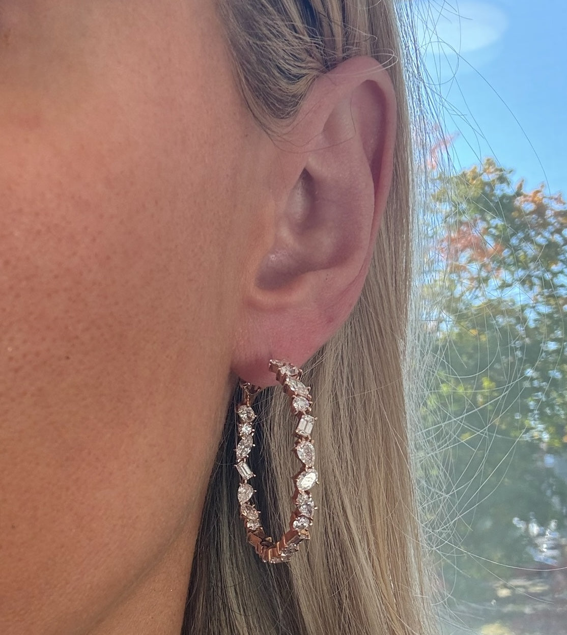 Close-up of a woman's ear wearing Mixed Shape Diamond Hoops; dazzling diamonds shine as her blonde hair is tucked behind her ear, with sunlight and blurred trees in the background.