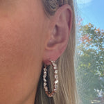 Close-up of a woman's ear wearing Mixed Shape Diamond Hoops; dazzling diamonds shine as her blonde hair is tucked behind her ear, with sunlight and blurred trees in the background.