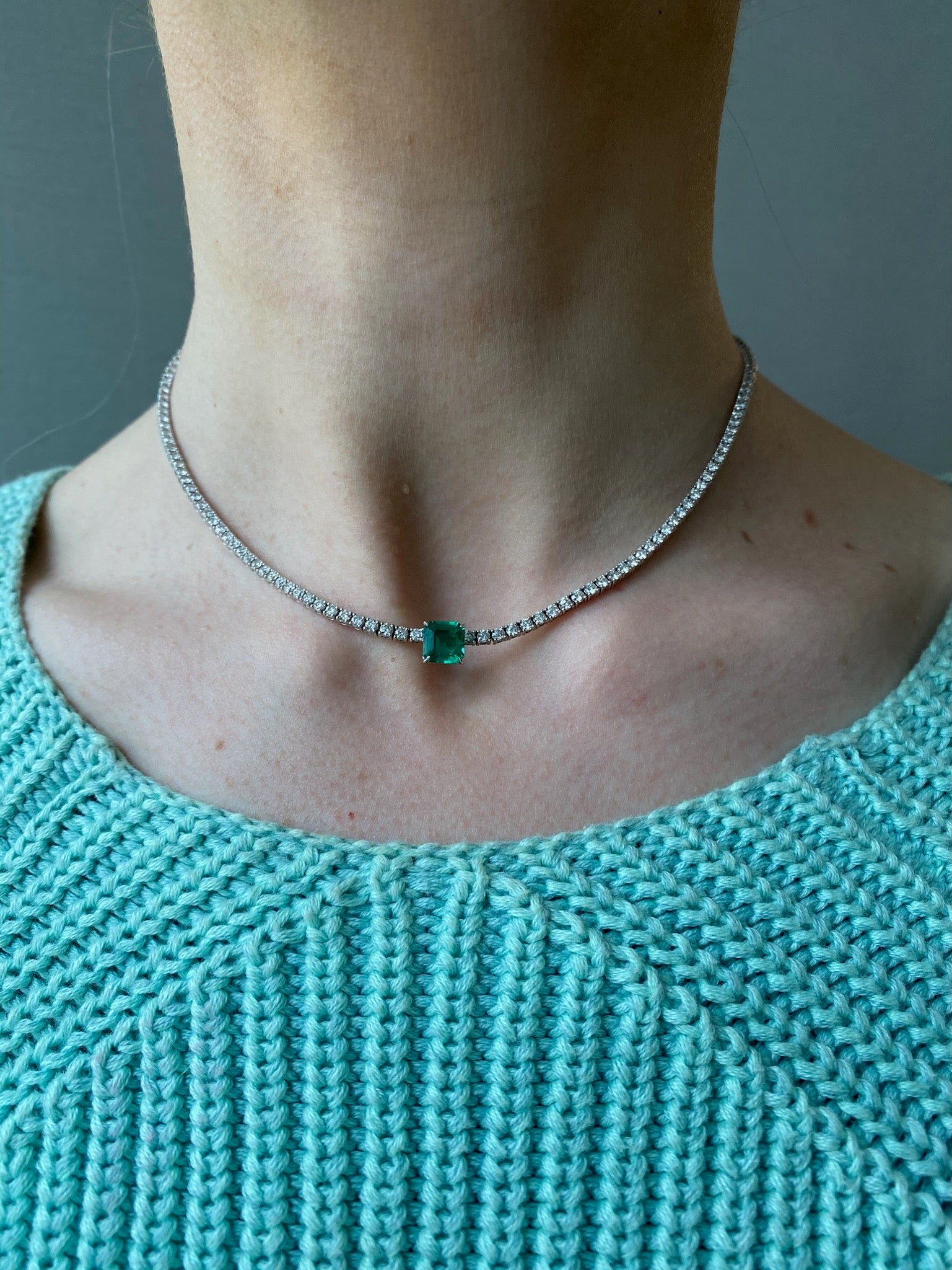 A close-up of a person wearing the Classic Tennis Necklace with Cushion Cut Emerald in Yellow Gold, paired with a light teal knitted top.