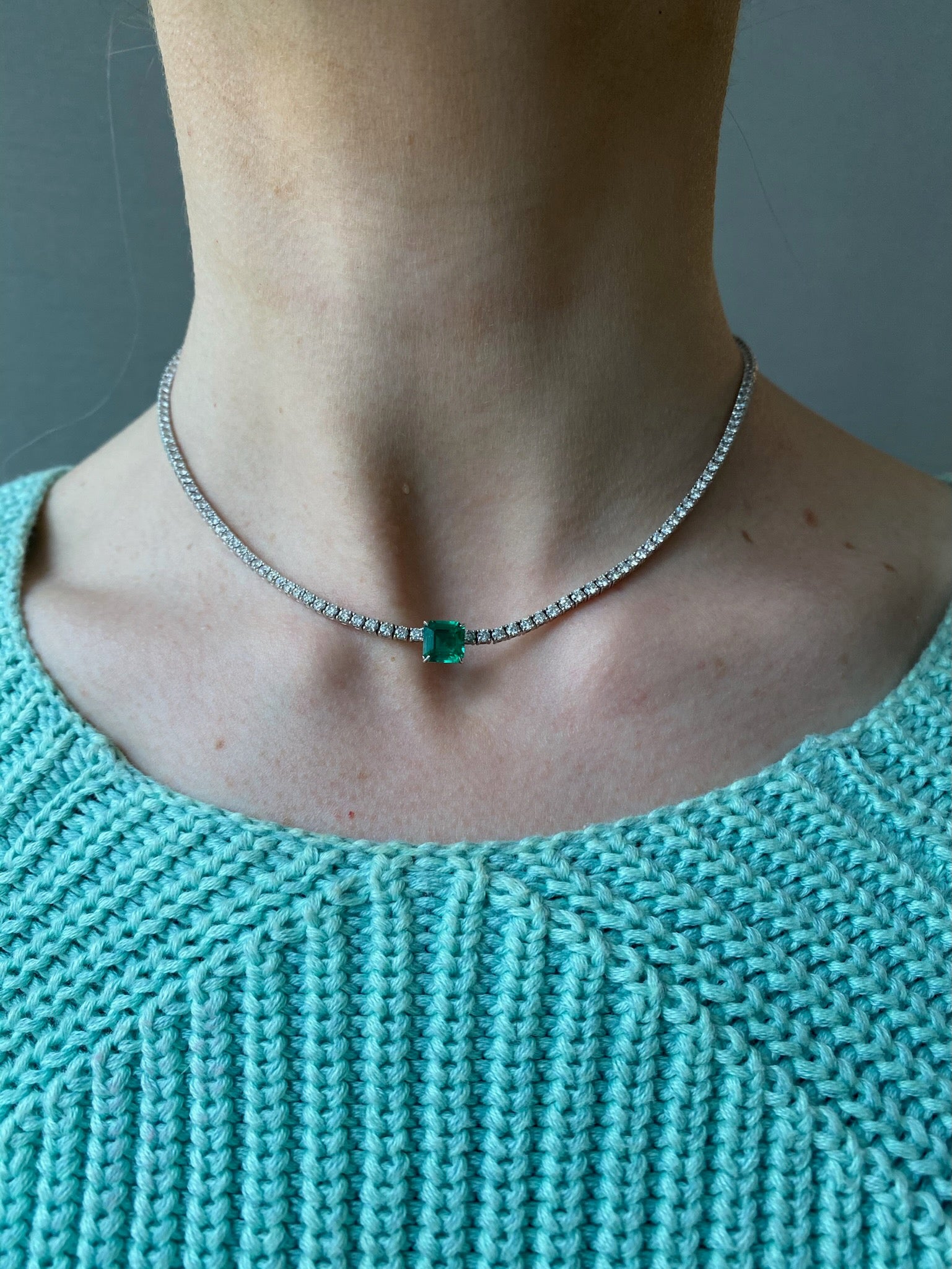 A person shown from shoulders to neck wears a light teal knitted top and the Classic Tennis Necklace with an Asscher Cut Emerald in White Gold.