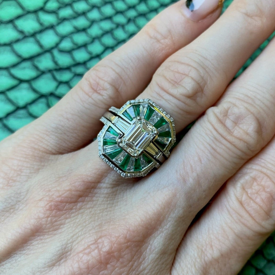 A manicured hand wears the Mosaic Tapered Baguette Emerald and Diamond Ring Jacket in white gold with a pavé diamond band, featuring baguette emeralds, diamonds, and a central rectangular stone against a textured green background.