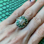 A manicured hand wears the Mosaic Tapered Baguette Emerald and Diamond Ring Jacket in white gold with a pavé diamond band, featuring baguette emeralds, diamonds, and a central rectangular stone against a textured green background.