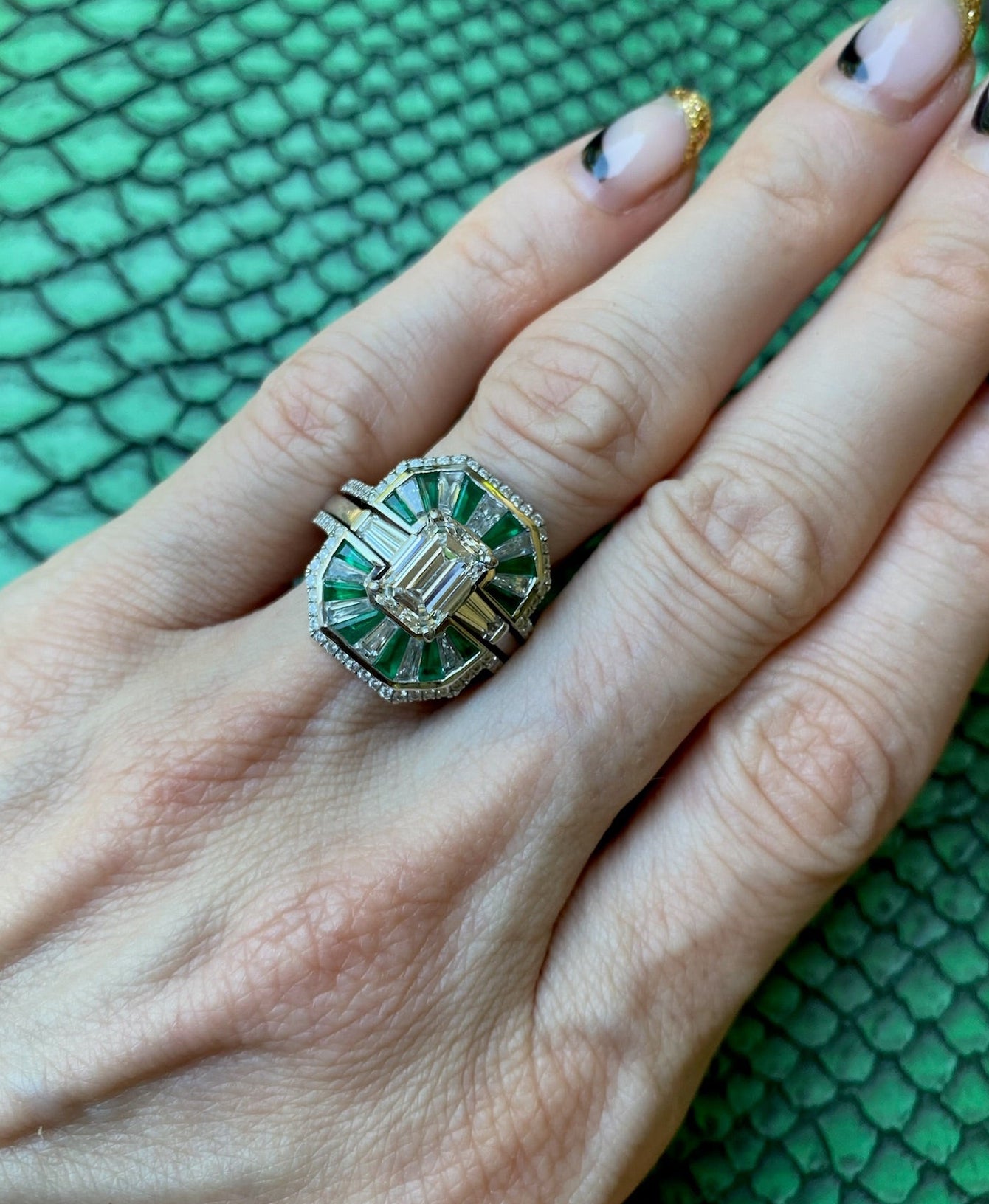 A manicured hand on a green surface wears the Bespoke Mosaic Tapered Baguette Emerald and Diamond Ring Jacket in White Gold, featuring a pavé diamond band, rectangular center diamond, baguette emeralds, and smaller diamonds.