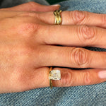 A hand wears a plain 18K yellow gold band on the middle finger and the Hover Pinky Ring in Yellow Gold with an Asscher Cut Diamond on the ring finger, both resting atop blue denim fabric.