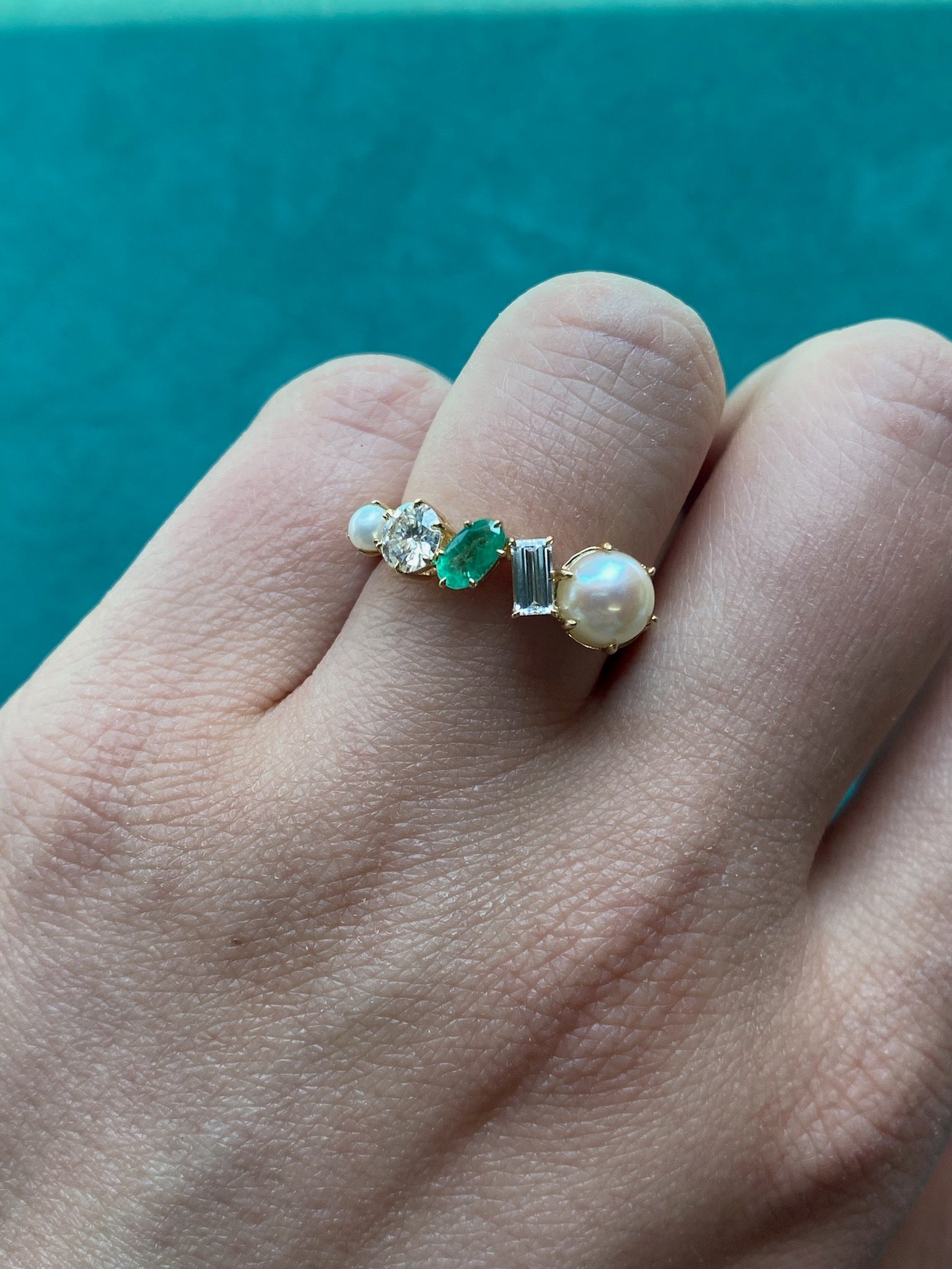 A hand wears the Mixed Shape Pearl, Diamond and Emerald Half-Eternity Band, featuring pearls, emeralds, diamonds, and small gemstones against a teal background.