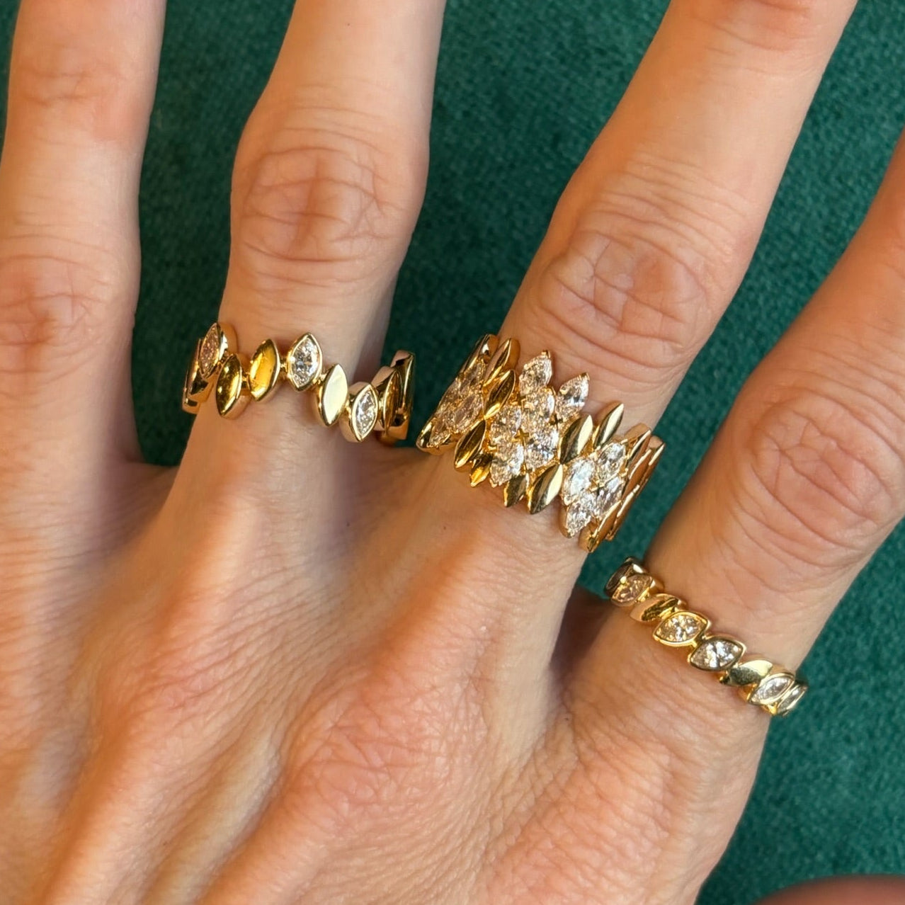A hand wears four Bezel Set Alternating Marquise Diamond Bands in yellow gold on the index, middle, ring, and pinky fingers, each featuring marquise-shaped diamonds in bezel settings against a green fabric background.
