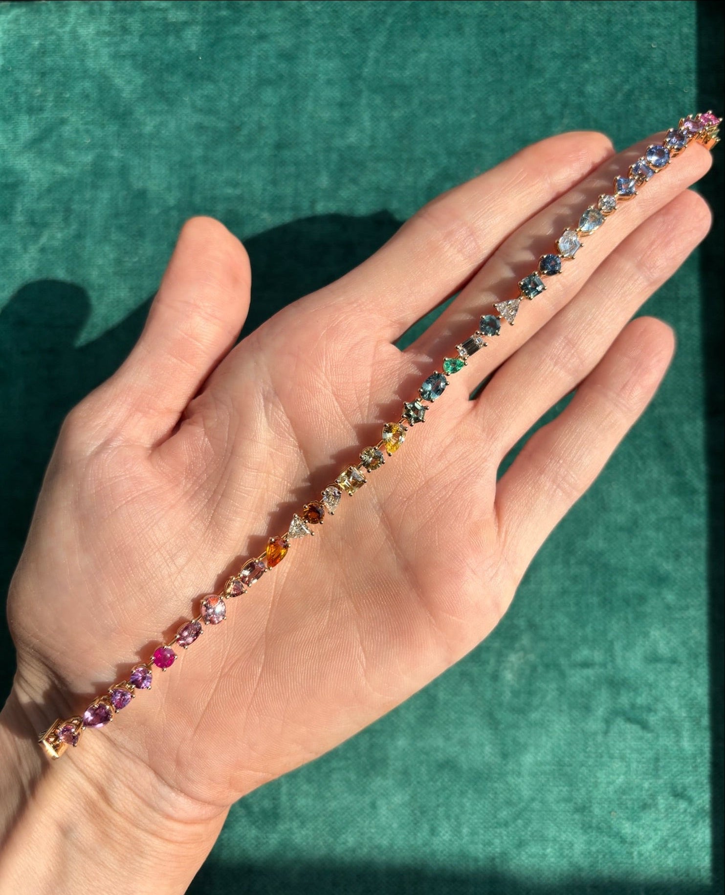 A hand holds the Bespoke Mixed Shape Multicolor Sapphire and Diamond Tennis Bracelet in yellow gold, its vibrant gems displayed in a line against a green textured background.