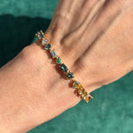 A close-up of a wrist wearing the Mixed Shape Multicolor Sapphire and Diamond Tennis Bracelet in yellow gold, featuring blue sapphires and multicolor diamonds, against a green background.