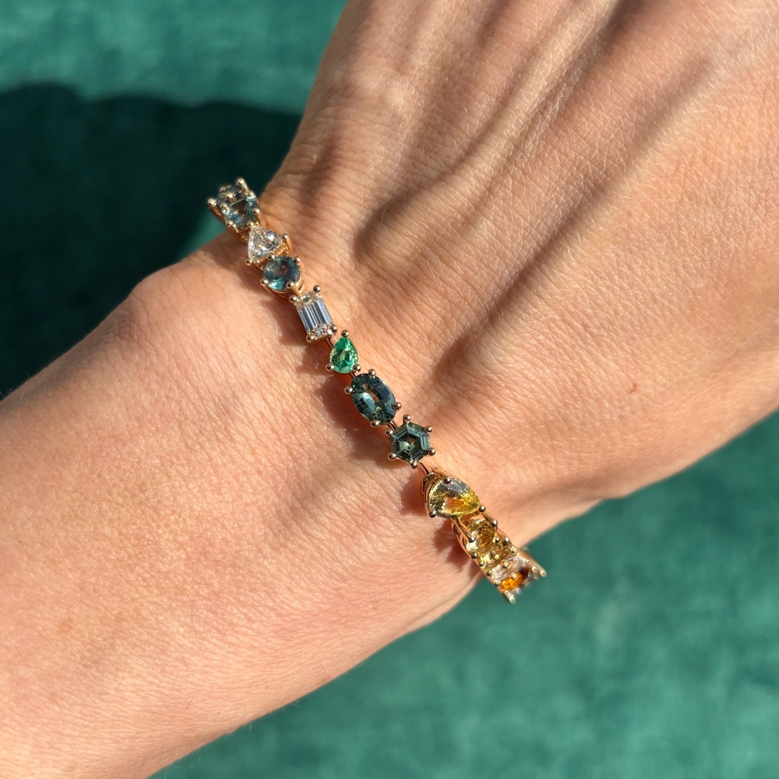 A close-up of a wrist wearing the Bespoke Mixed Shape Multicolor Sapphire and Diamond Tennis Bracelet in Yellow Gold, showcasing vibrant sapphires and diamonds of various shapes against a green background.