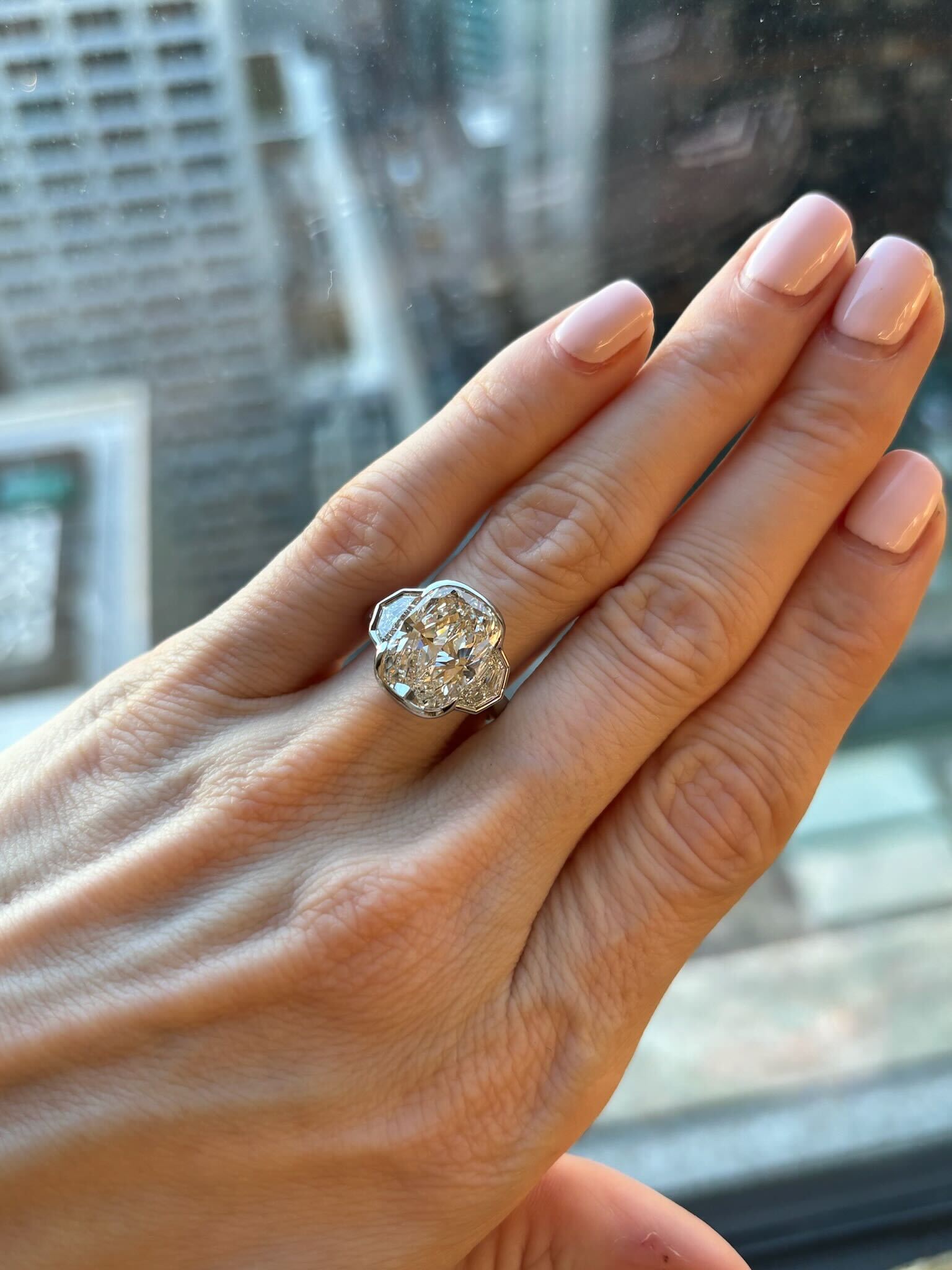 A hand with neatly manicured, pale pink nails wears the Radiant Cut Diamond Engagement Ring with Bezel Setting and Epaulette Side Stones—an elegant silver piece that sparkles against the blurred cityscape through the window.