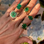 A manicured hand displays five gold rings, each with vibrant green emeralds in unique shapes and settings, including the Mosaic Asscher Cut Emerald and Baguette Diamond Cocktail Ring in Platinum. Sunlight highlights the gems against a blurred floral background.
