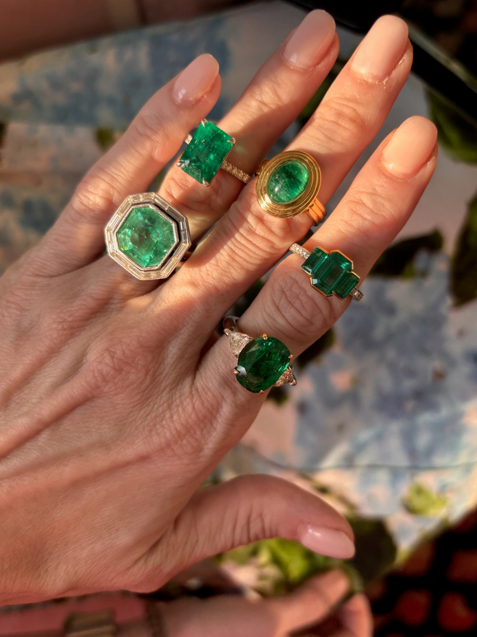 A manicured hand displays five gold rings, each with vibrant green emeralds in unique shapes and settings, including the Mosaic Asscher Cut Emerald and Baguette Diamond Cocktail Ring in Platinum. Sunlight highlights the gems against a blurred floral background.