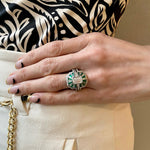 A hand with the Mosaic Tapered Baguette Emerald and Diamond Ring Jacket in White Gold with Pavé Diamond Band rests on white pants. The look is finished with a gold waist chain, minimal black nails, and a black-and-white patterned top.