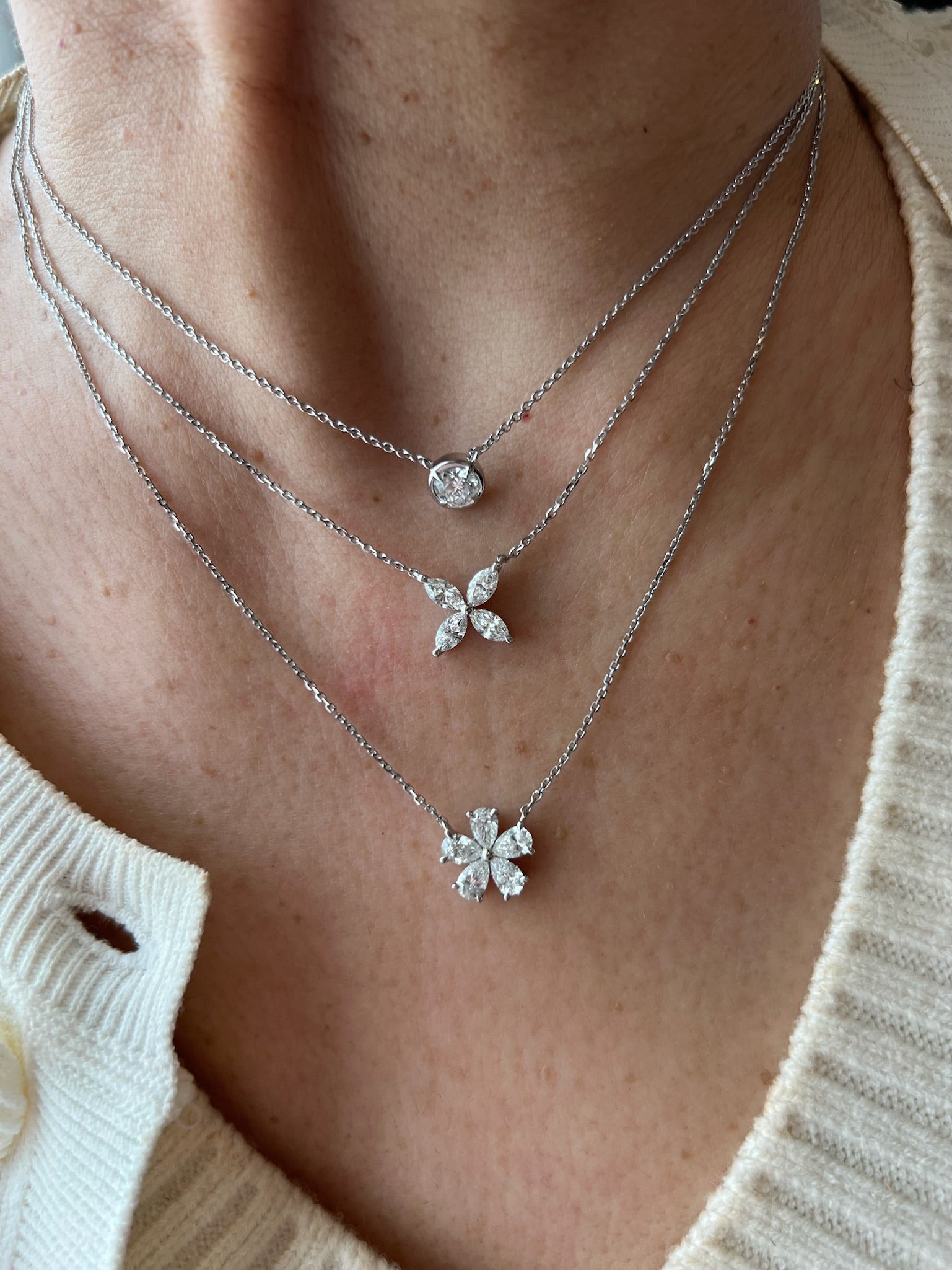 A close-up of a person in a cream knit top wearing three layered silver necklaces with diamond pendants, featuring the Bezel Set Marquise Diamond Floral Pendant: a single marquise, a four-petal cluster, and a larger floral shape.