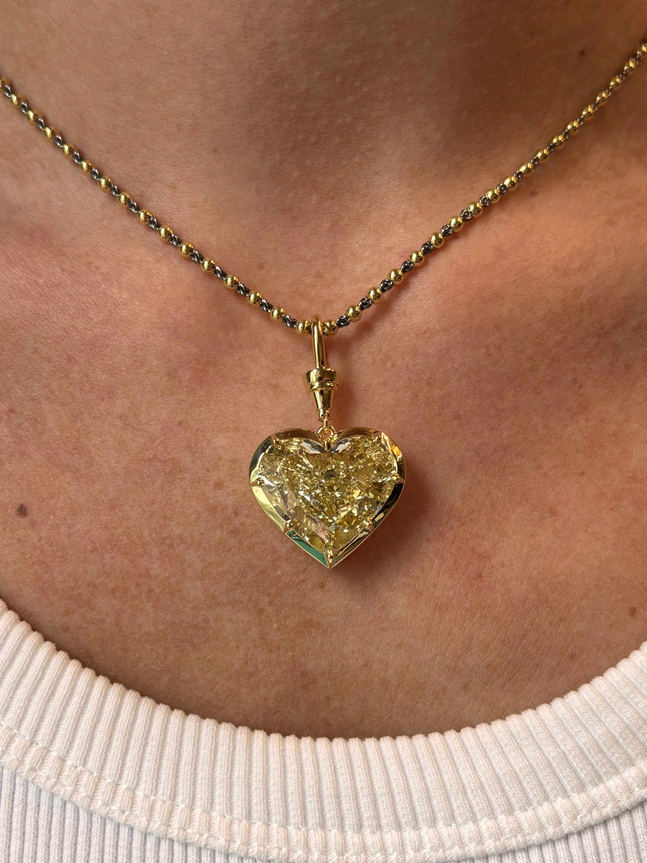 A close-up of a person wearing the Collet Set Heart Shape Fancy Yellow Diamond Necklace in Yellow Gold, featuring an 18k yellow gold chain and a heart-shaped yellow diamond pendant, against their skin and a ribbed white top.