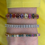 A close-up of a wrist showcases the Multicolor Oval Sapphire Tennis Bracelet, sparkling with vibrant sapphires and set in luxurious 18K yellow gold above a yellow sleeve.