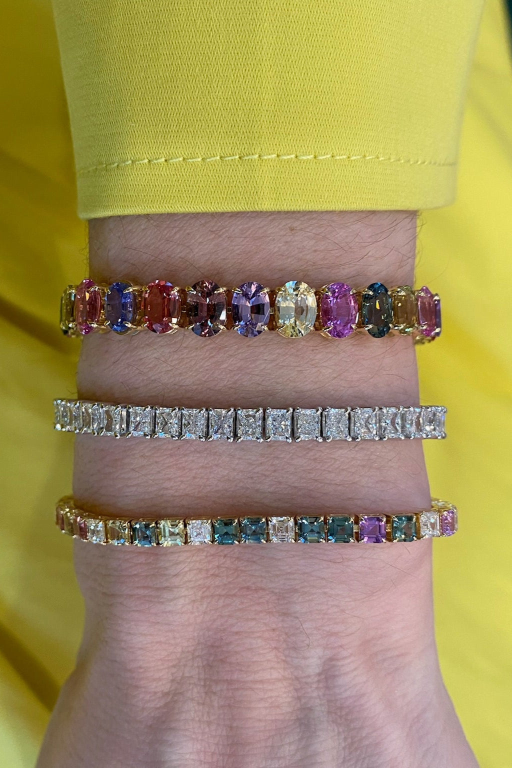 A close-up of a wrist showcases the Multicolor Oval Sapphire Tennis Bracelet, sparkling with vibrant sapphires and set in luxurious 18K yellow gold above a yellow sleeve.