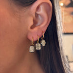 Close-up of a person’s ear wearing the Collet Set Emerald Cut Diamond Drop Earring in Black Rhodium and Yellow Gold. Their straight brown hair frames the elegant gold hoop with a large emerald-cut diamond drop.