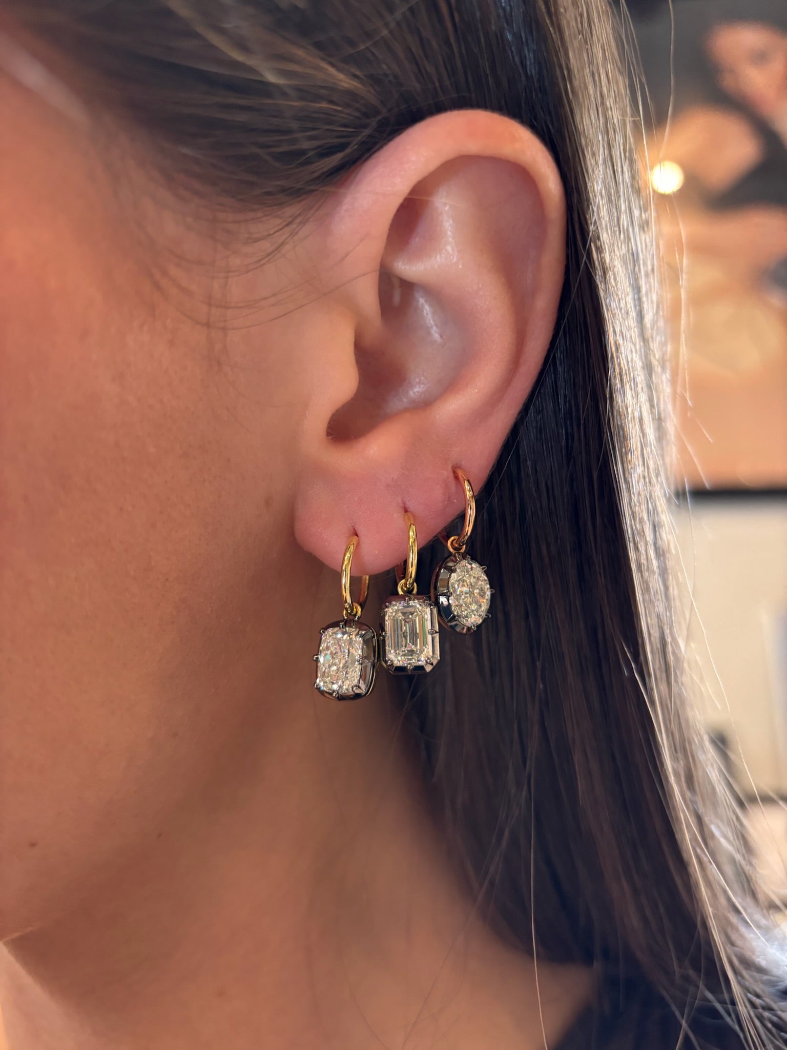 Close-up of a person’s ear wearing the Collet Set Emerald Cut Diamond Drop Earring in Black Rhodium and Yellow Gold. Their straight brown hair frames the elegant gold hoop with a large emerald-cut diamond drop.
