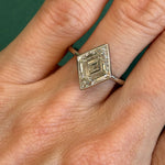 A hand wearing the Bezel Set Lozenge Diamond Engagement Ring, crafted in 18K white gold with a large, geometric lozenge-shaped diamond, is displayed against a green background.