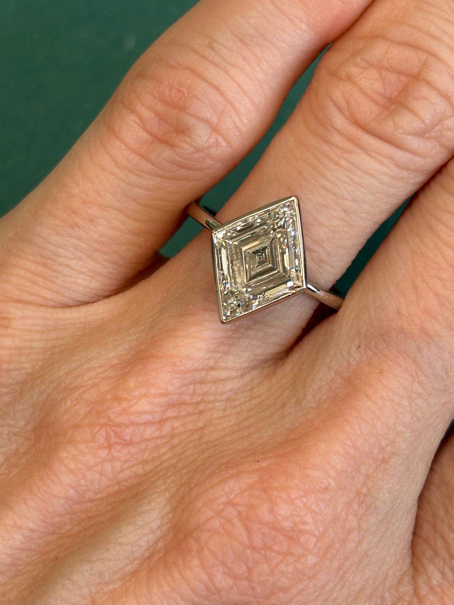 A hand wearing the Bezel Set Lozenge Diamond Engagement Ring, crafted in 18K white gold with a large, geometric lozenge-shaped diamond, is displayed against a green background.