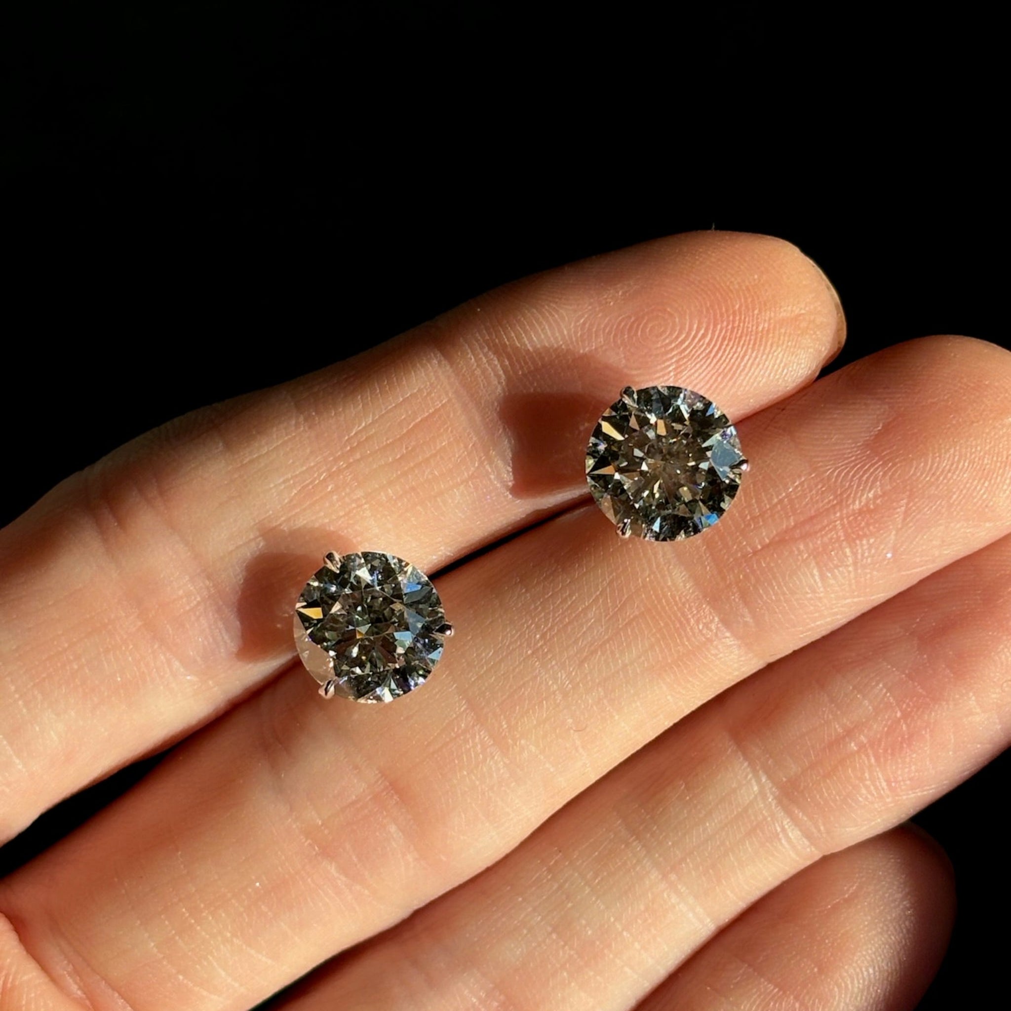 A close-up of a hand holding Classic Round Diamond Studs in White Gold, featuring two round brilliant diamonds set in a martini setting against a dark background.