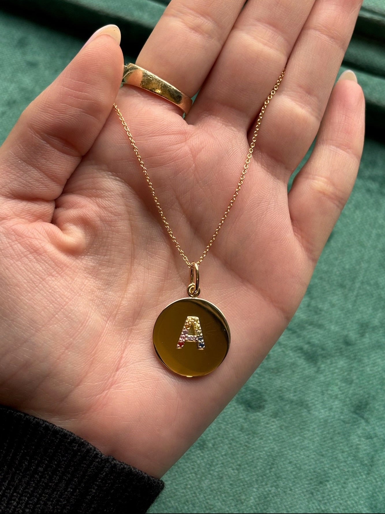 A hand holds a Round Pendant Charm in yellow gold featuring a letter motif in rainbow sapphires, ideal for customizable jewelry fans. A gold ring is also worn, with both pieces set against a green fabric backdrop.