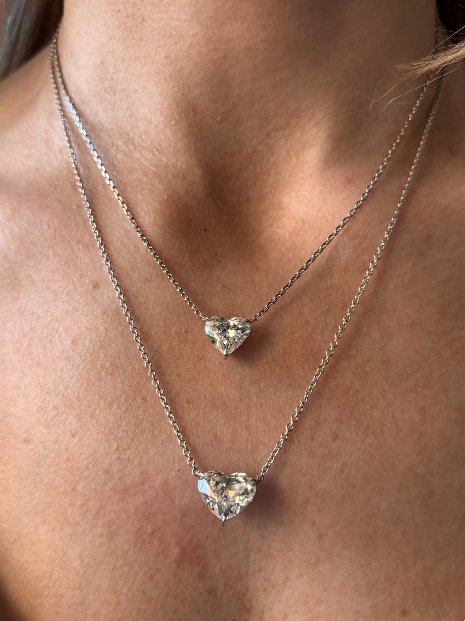 Close-up of a person's neck wearing two Heart Diamond Solitaire Pendant necklaces, each featuring a sparkling heart-shaped diamond solitaire, with one necklace slightly shorter than the other.