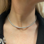 A close-up of a woman’s neck showcases the Large Adjustable Round Brilliant Diamond Tennis Necklace in Yellow Gold, paired with part of a black blazer and a decorative brooch visible on the left.