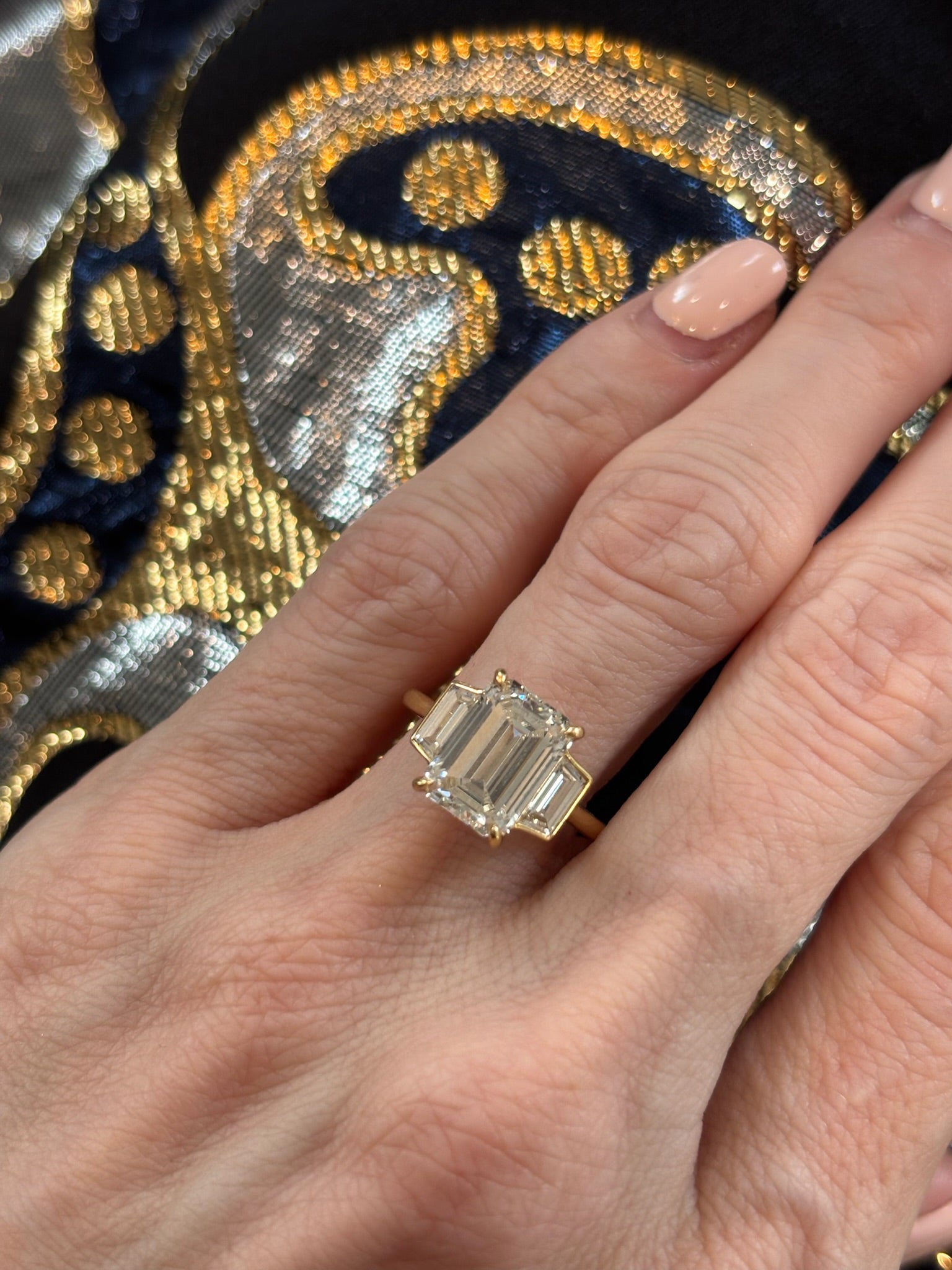 A hand with manicured nails wears the Emerald Cut Diamond Engagement Ring with Trapezoid Side Stones, crafted in 18k yellow gold and set with three emerald-cut diamonds. The backdrop displays patterned gold, navy blue, and silver embroidery.