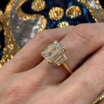 A close-up of a hand wearing the Emerald Cut Diamond Engagement Ring with Trapezoid Side Stones, set in 18k yellow gold, shown against detailed blue and gold embroidery.