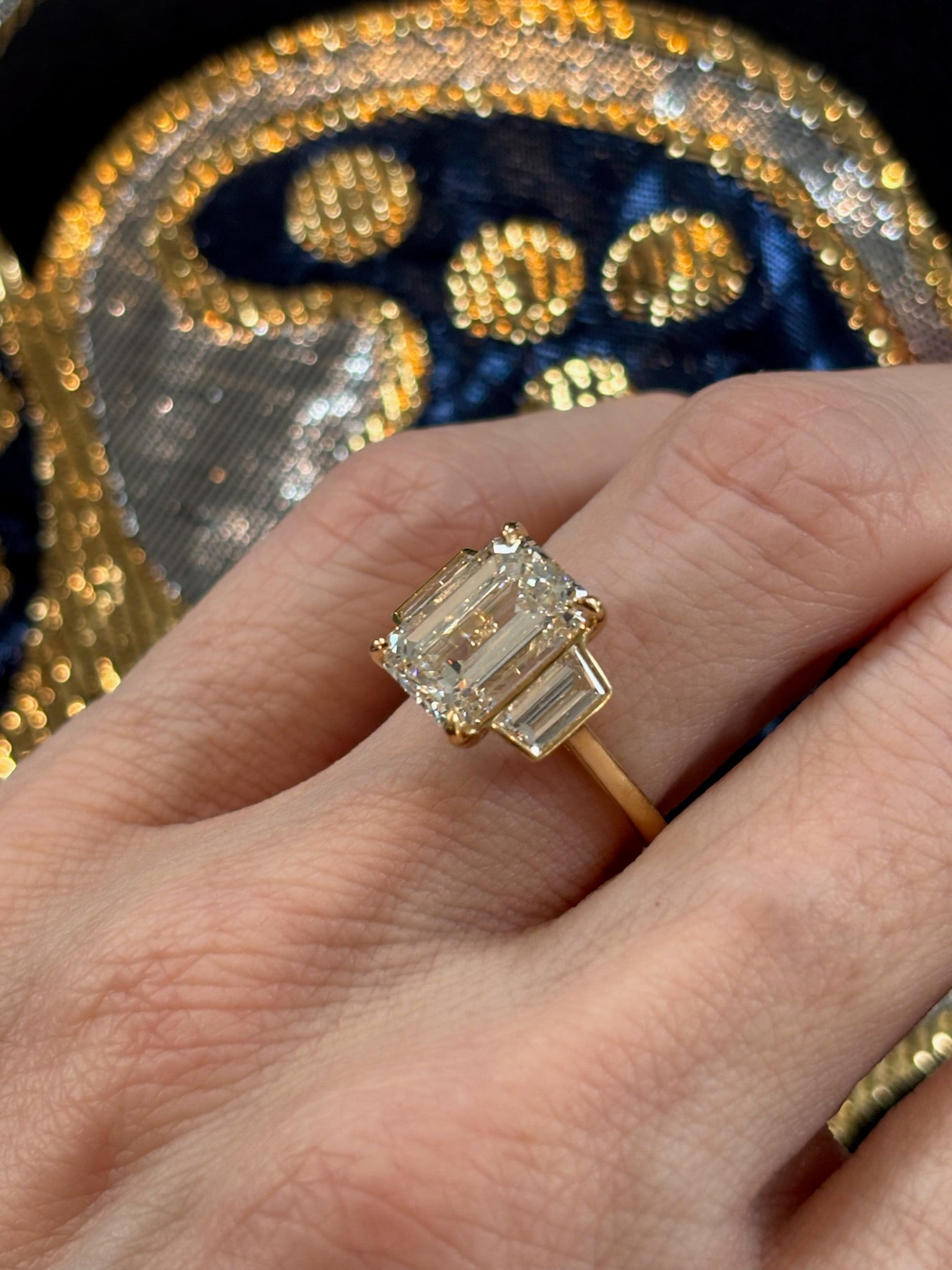 A close-up of a hand wearing the Emerald Cut Diamond Engagement Ring with Trapezoid Side Stones, set in 18k yellow gold, shown against detailed blue and gold embroidery.