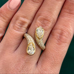A hand showcases the Snake Open Space Ring in yellow gold with two pear shape diamonds, each surrounded by pavé diamonds, set against a dark green background.
