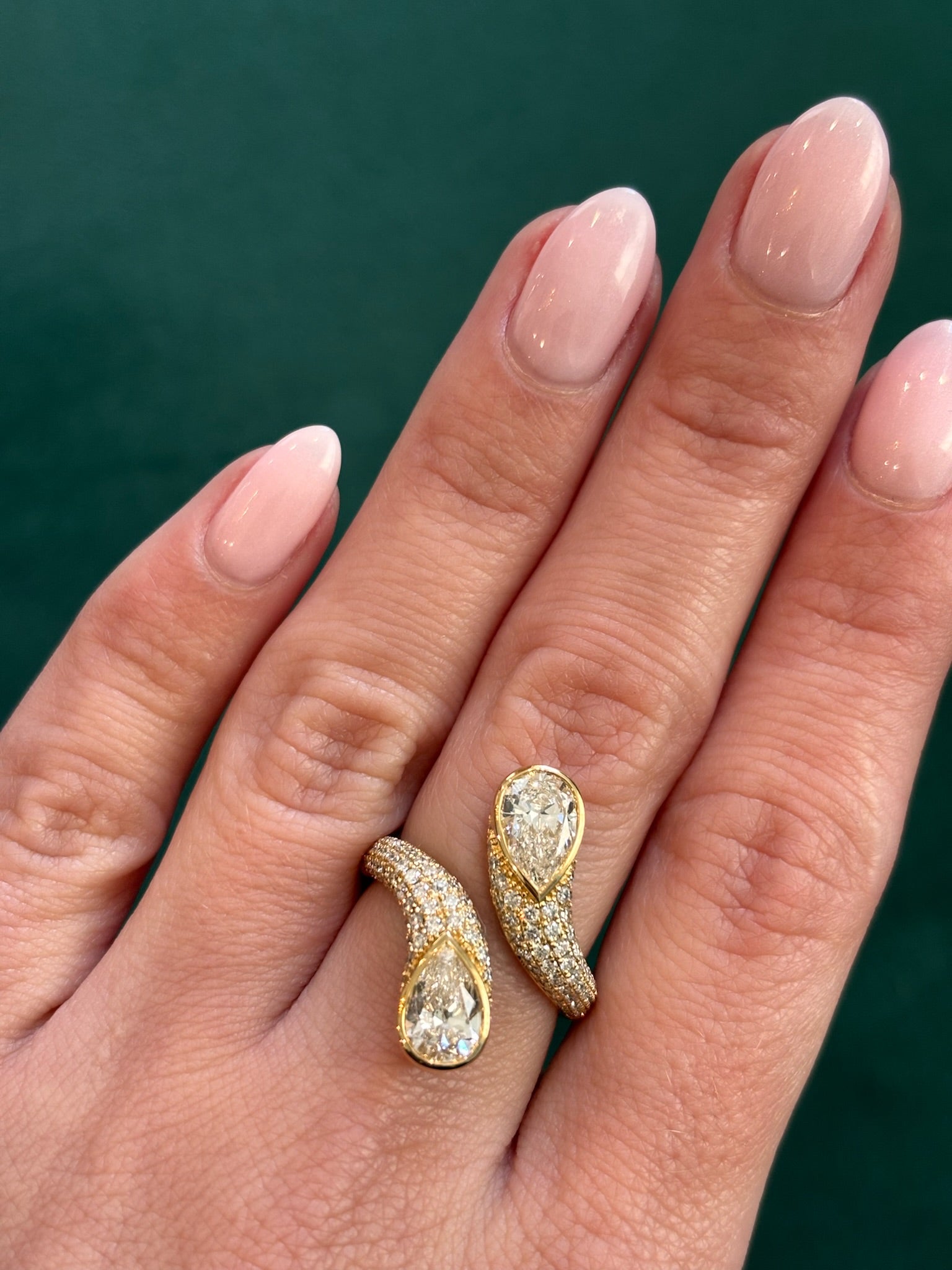 A hand with light pink, almond-shaped nails wears the Snake Open Space Ring in yellow gold, featuring two facing pear shape diamonds encircled by pavé on a solid dark green background.