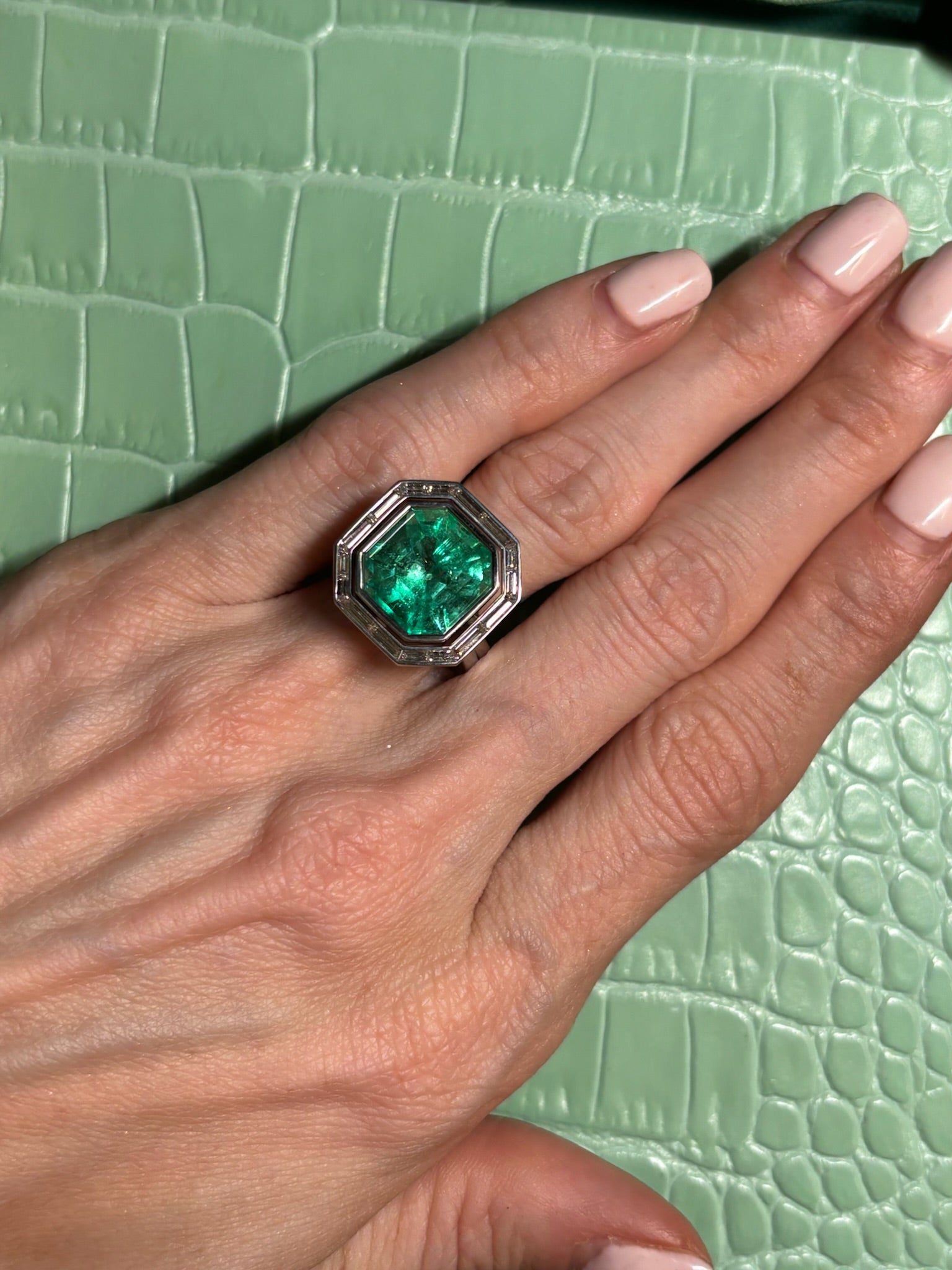 A hand with light pink manicured nails wears the Mosaic Asscher Cut Emerald and Baguette Diamond Cocktail Ring in Platinum, resting on a textured light green crocodile leather-like surface.