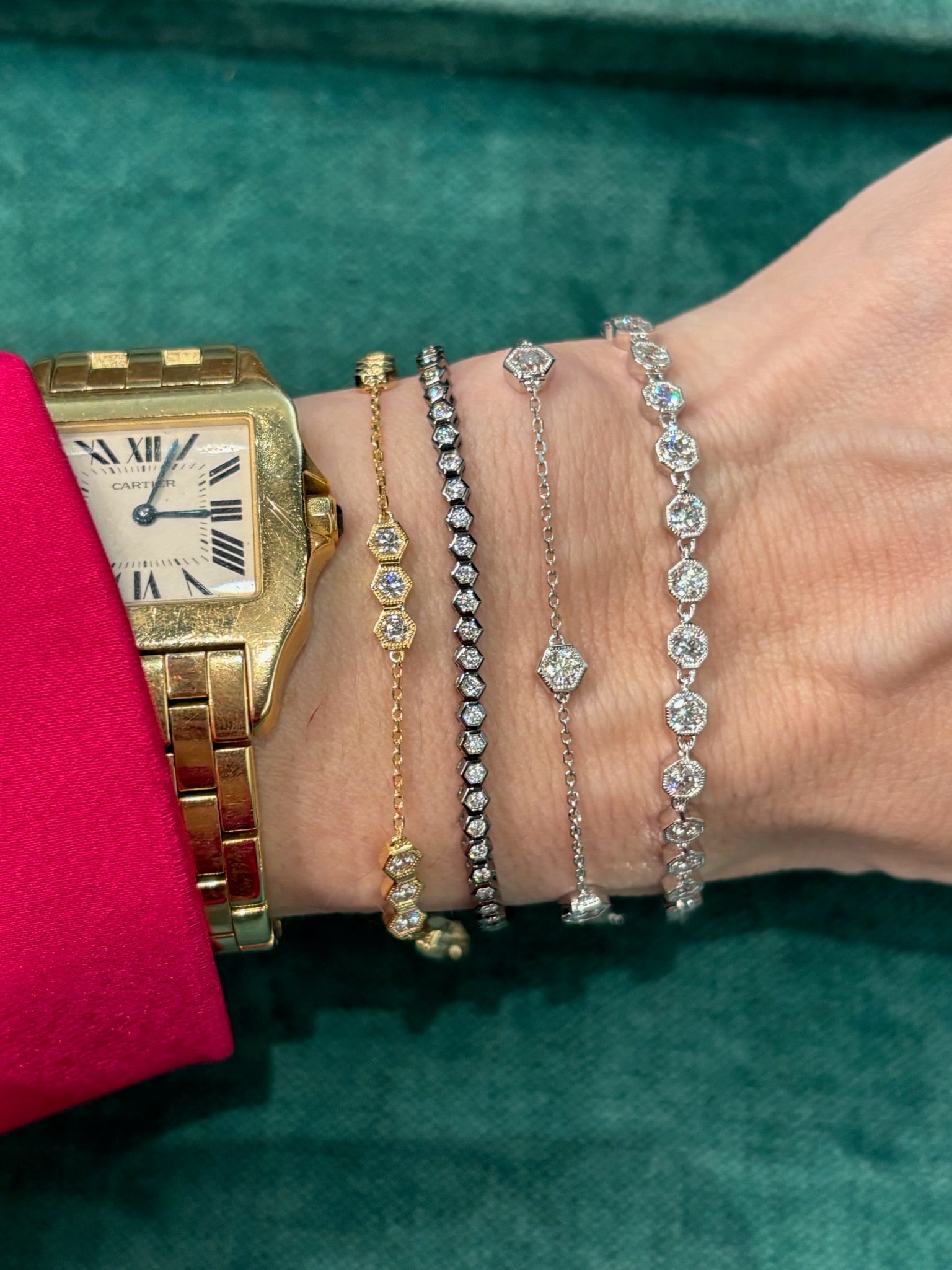 A wrist adorned with a gold Cartier watch, two gold bracelets—including the Serpentine Station Bracelet with Hexagon Bezel Set Diamonds—and a silver hexagon bracelet rests on green, with the wearer in a red sleeve.