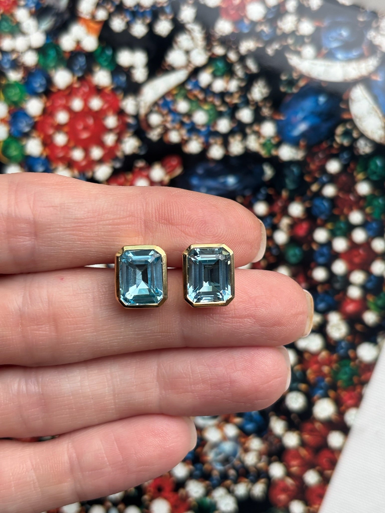 A hand displays Bezel Set Emerald Cut Aquamarine Stud Earrings in Yellow Gold against a vibrant, jeweled background.