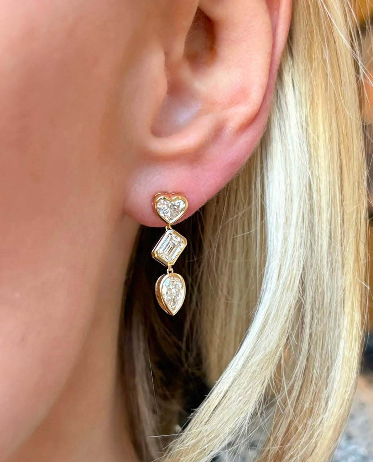 A close-up shows a woman with light blonde hair wearing Bezel Set Mixed Shape Diamond Drop Earrings in 18K gold, featuring heart, rectangle, and teardrop-shaped diamonds arranged from top to bottom.