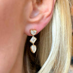 A close-up shows a woman with light blonde hair wearing Bezel Set Mixed Shape Diamond Drop Earrings in 18K gold, featuring heart, rectangle, and teardrop-shaped diamonds arranged from top to bottom.