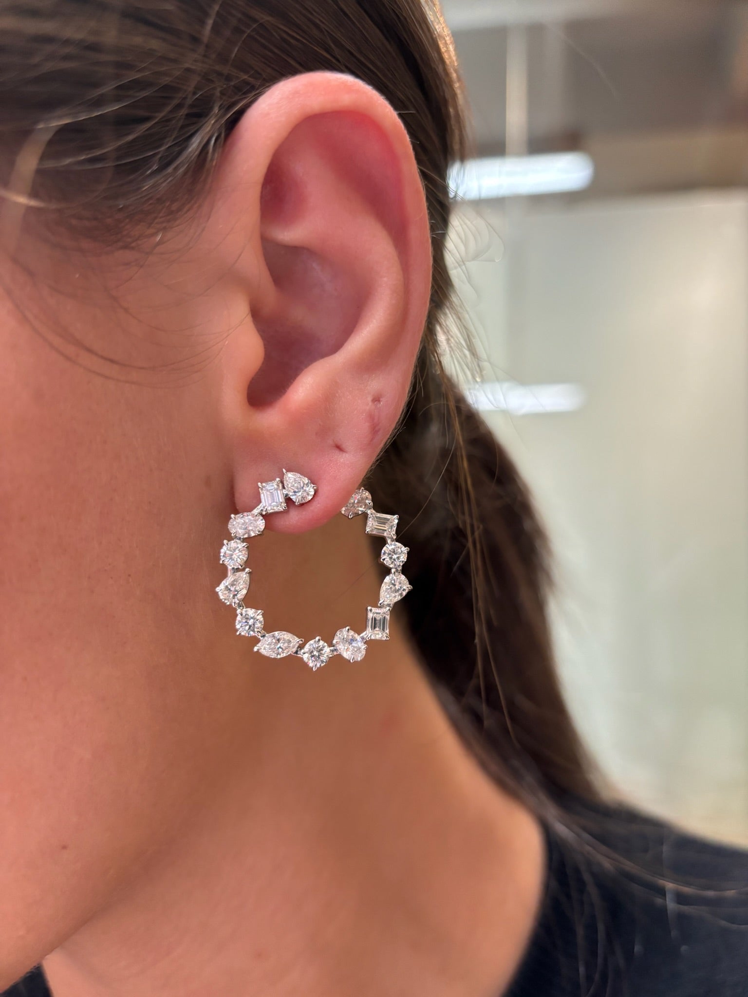 Close-up of a person’s ear wearing Mixed Shape Diamond Front to Back Hoops in White Gold, featuring sparkling clear gemstones. The blurred neutral background highlights the exquisite shine of these diamond hoops.