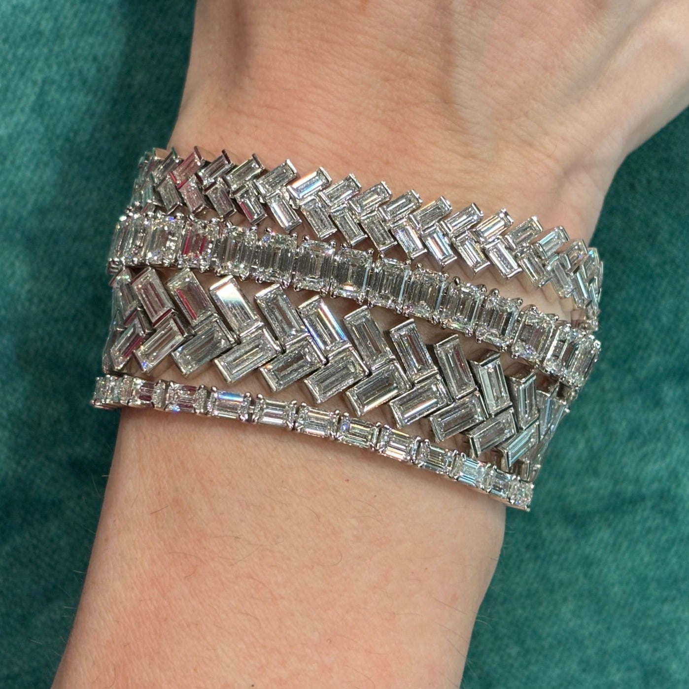 A close-up of a wrist wearing three Half-Bezel Set Chevron Diamond Tennis Bracelets in white gold, each with baguette diamonds in geometric zigzag patterns, set against a textured green fabric background.