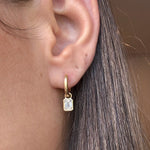 Close-up of a person's ear wearing Huggies with Bezel Set Emerald Cut Diamond Charms in Yellow Gold from Serpentine Jewels. Their straight brown hair frames the earring—customize your own pair for a unique look.