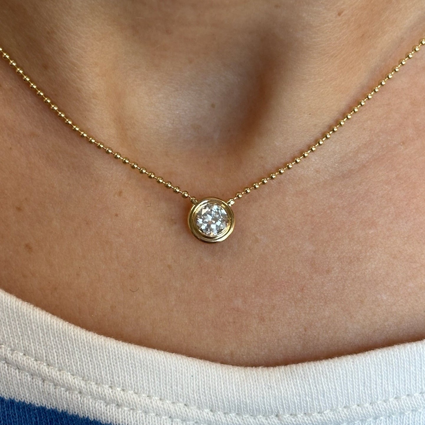A close-up of the Bezel Set Round Diamond Ball Chain Necklace in Yellow Gold, featuring a round diamond pendant, worn around the neck with a white shirt with a blue stripe.