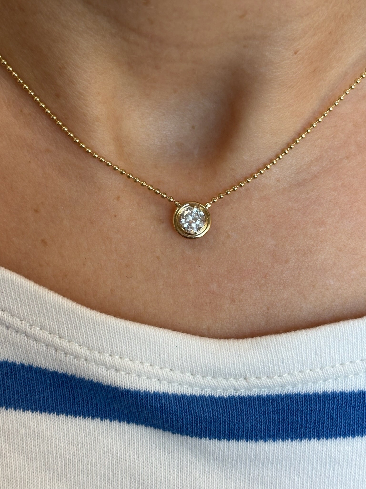 A close-up of the Bezel Set Round Diamond Ball Chain Necklace in Yellow Gold, featuring a round diamond pendant, worn around the neck with a white shirt with a blue stripe.