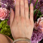 A hand with light purple nail polish wears the Chevron Baguette Diamond Bangle in White Gold, surrounded by pink roses, purple lilacs, and green leaves against a backdrop of vibrant blooming flowers.